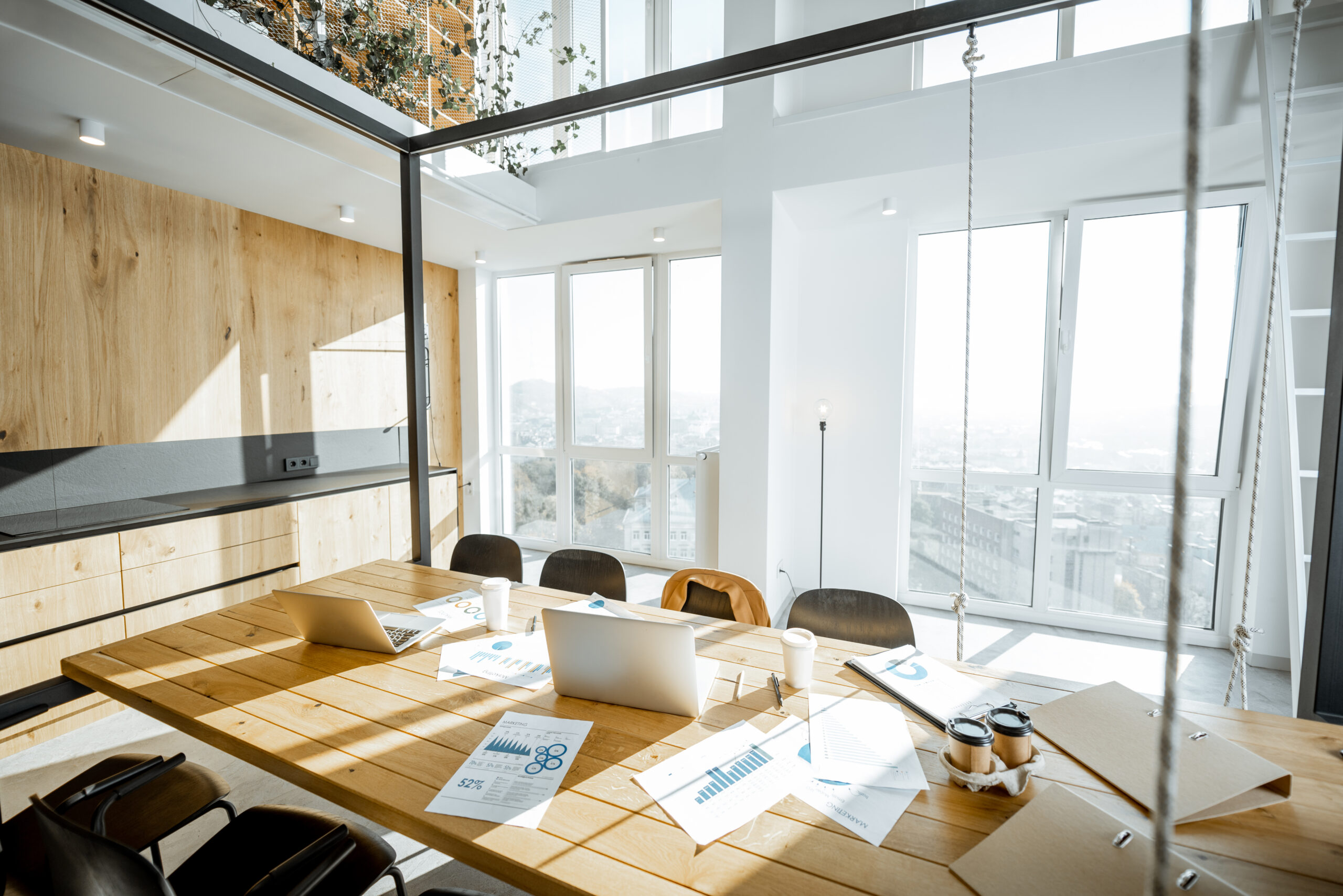 Arbeitsplatz mit grossem Holztisch und Büromaterial, ohne Personen, in einem hellen, sonnigen Büroraum, weitläufiger Blick ins Innere